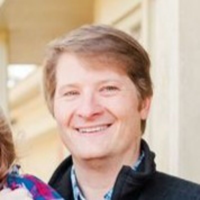 The image shows a man with fair skin and light brown hair. He is smiling and appears to be in an outdoor setting. He is wearing a dark jacket over a patterned shirt. The background is blurred, suggesting a shallow depth of field.